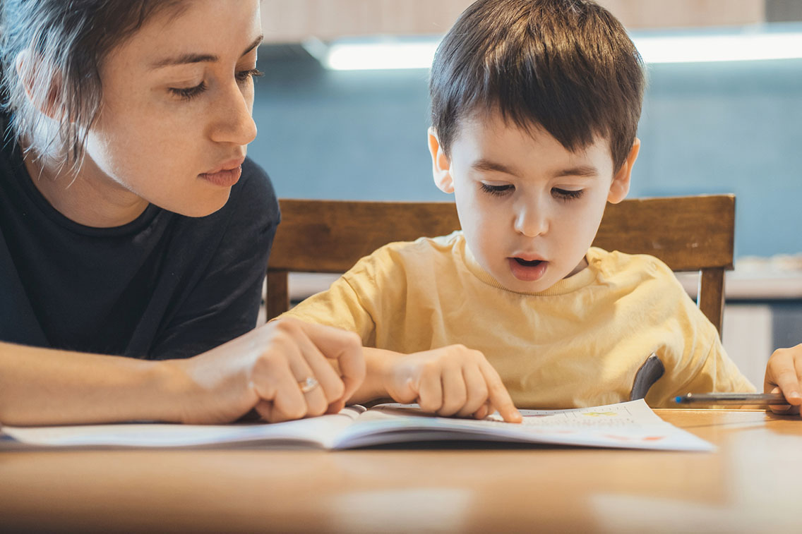 Little boy with autism and his mother sitting together at table and reading book